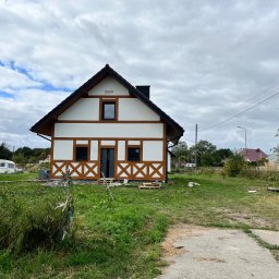 Max Construction - Dwukondygnacyjny dom z białymi ścianami i brązowymi belkami w stylu szachulcowym, z czarnym dachem i numerem '2024' na szczycie, otoczony zielenią.