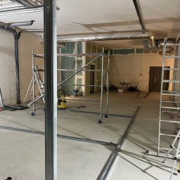 Max Construction - Wnętrze w trakcie prac wykończeniowych: widoczne rusztowania, profile pod płyty gipsowe na podłodze i ścianach, częściowo zamontowane płyty GK, instalacje pod sufitem.