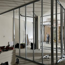 Max Construction - Montaż metalowej konstrukcji ścian działowych wewnątrz pomieszczenia, widoczne płyty gipsowe i narzędzia, dwóch mężczyzn przy pracy.