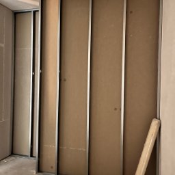 Max Construction - Montaż ścianki działowej z płyty kartonowo-gipsowej na metalowym stelażu, widoczny szlifierka kątowa i zwinięty materiał izolacyjny.