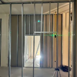 Montaż ścianek działowych z płyt karton-gips na metalowym stelażu, widoczny laser krzyżowy, drabina w przejściu. Etap prac wykończeniowych wnętrz.