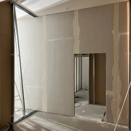 Max Construction - Wnętrze w trakcie prac wykończeniowych: ściany z płyt gipsowo-kartonowych, widoczne metalowe profile i szpachlowane łączenia. Przejście do kolejnego pomieszczenia.