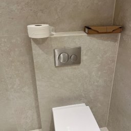 Max Construction - Minimalistyczna toaleta w jasnych płytkach z ukrytym systemem spłukiwania Geberit, papierem toaletowym i kartonem na półce. Nowoczesne wykończenie wnętrza.