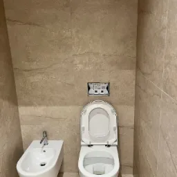 Beżowa łazienka z płytkami imitującymi kamień: biała toaleta z otwartą deską i bidet, chromowana armatura, minimalistyczny design.