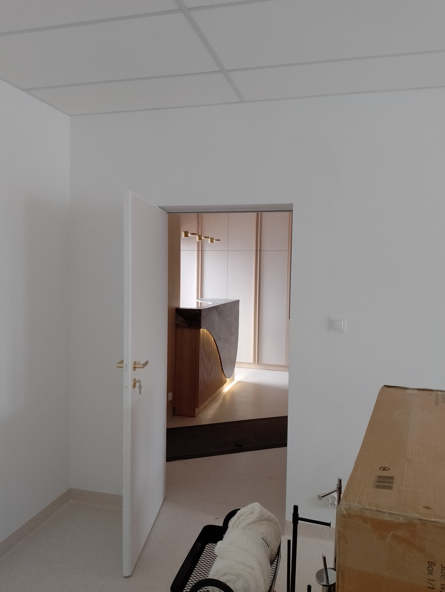 Widok z korytarza na recepcję z marmurowym blatem i podświetlanymi panelami w nowym biurze, widoczne białe ściany i sufit podwieszany.