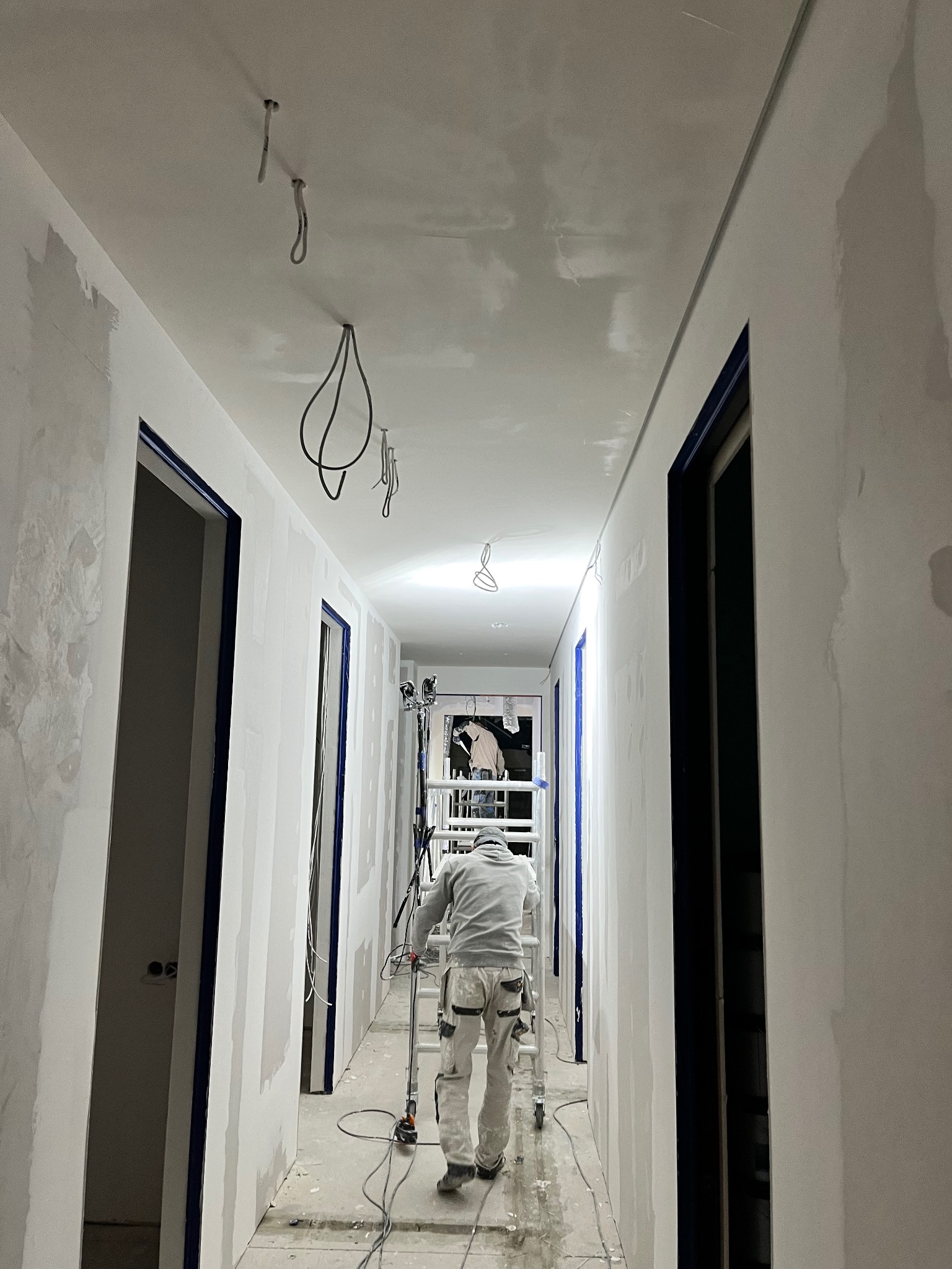 Korytarz w trakcie szpachlowania ścian i sufitu, widoczny pracownik na rusztowaniu i przewody elektryczne zwisające z sufitu. Przygotowanie do malowania.