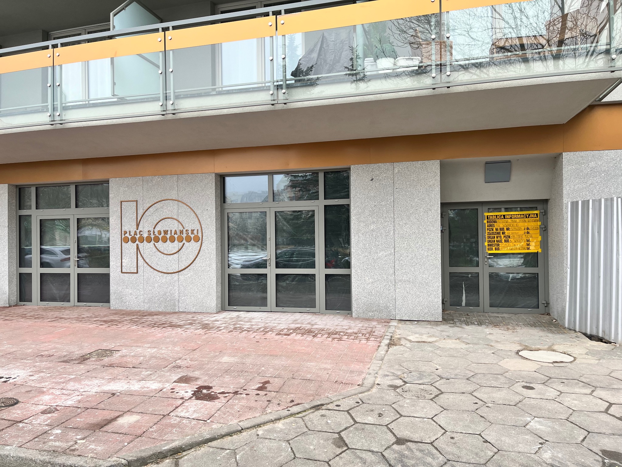 Szara fasada budynku z napisem 'Plac Słowiański 10' i żółtą tablicą informacyjną przy wejściu. Widok na fragment balkonu z przeszkloną balustradą.