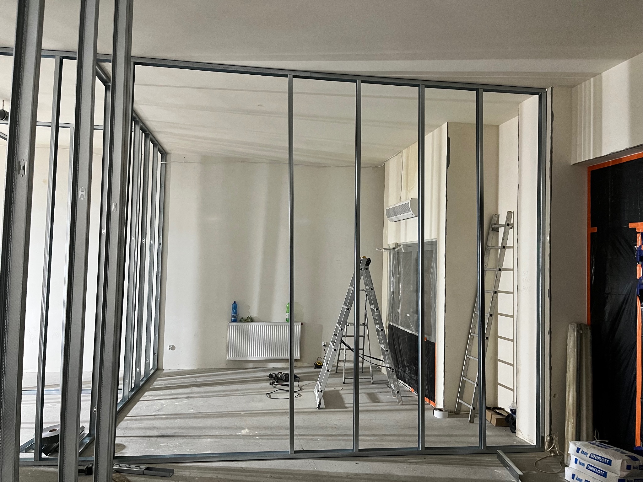 Montaż metalowego szkieletu ścian działowych w surowym wnętrzu. Widoczne profile, drabiny, narzędzia i worki z materiałem budowlanym. Prace wykończeniowe w toku.