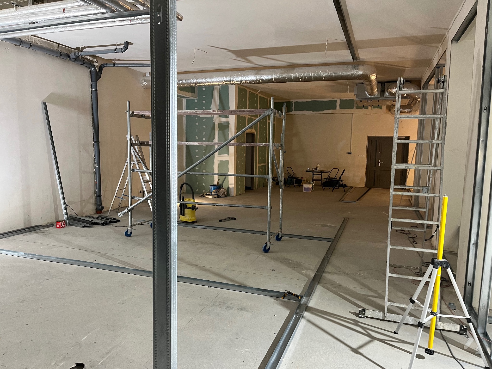 Wnętrze w trakcie prac wykończeniowych: widoczne rusztowania, profile pod płyty gipsowe na podłodze i ścianach, częściowo zamontowane płyty GK, instalacje pod sufitem.