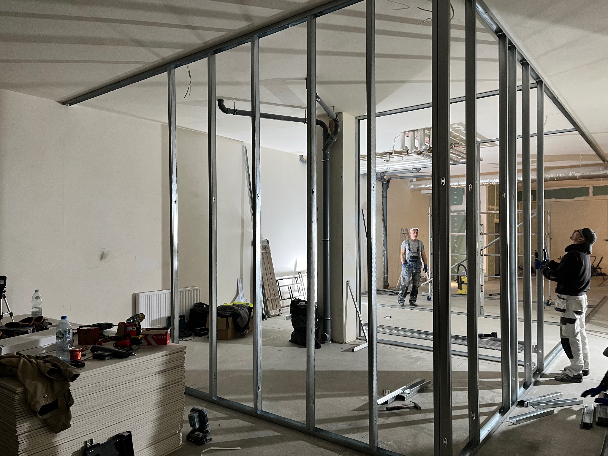 Montaż metalowej konstrukcji ścian działowych wewnątrz pomieszczenia, widoczne płyty gipsowe i narzędzia, dwóch mężczyzn przy pracy.