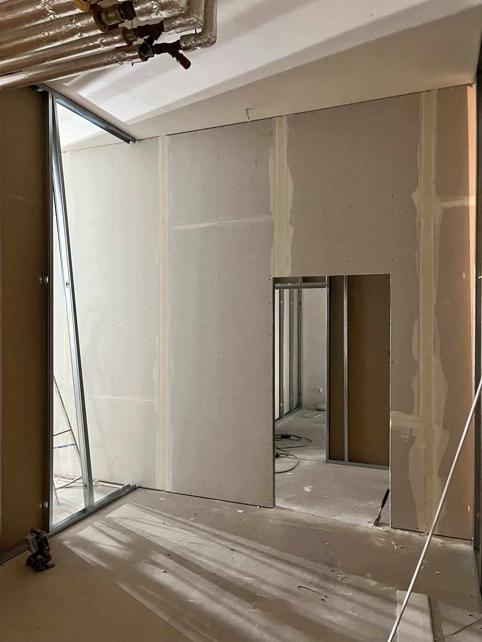 Wnętrze w trakcie prac wykończeniowych: ściany z płyt gipsowo-kartonowych, widoczne metalowe profile i szpachlowane łączenia. Przejście do kolejnego pomieszczenia.