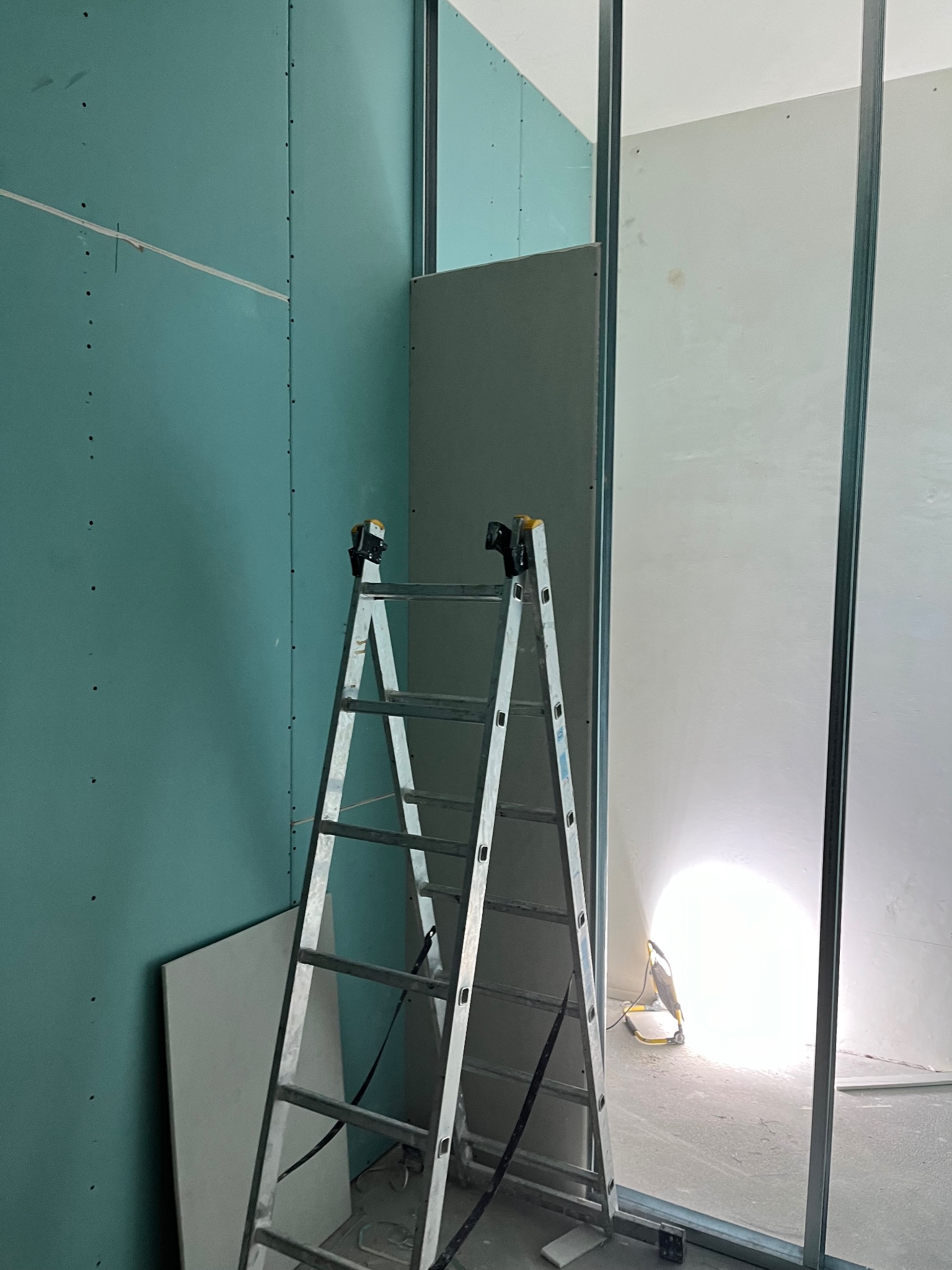 Montaż płyt kartonowo-gipsowych na metalowym stelażu, widoczna drabina aluminiowa i fragment oświetlenia budowlanego. Ściany w kolorze szarozielonym.