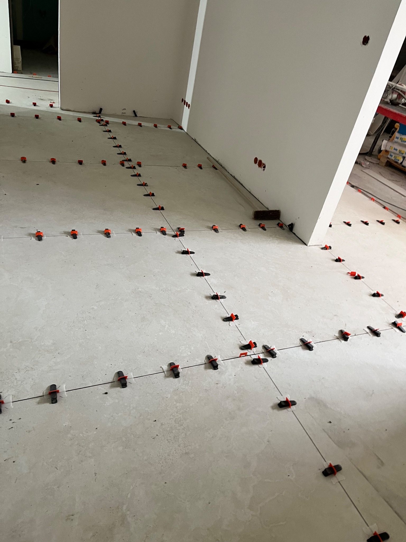 Ułożone płytki podłogowe z systemem poziomowania, widoczne fugi i elementy mocujące. W tle fragment ściany i pomieszczenia w trakcie wykończenia.