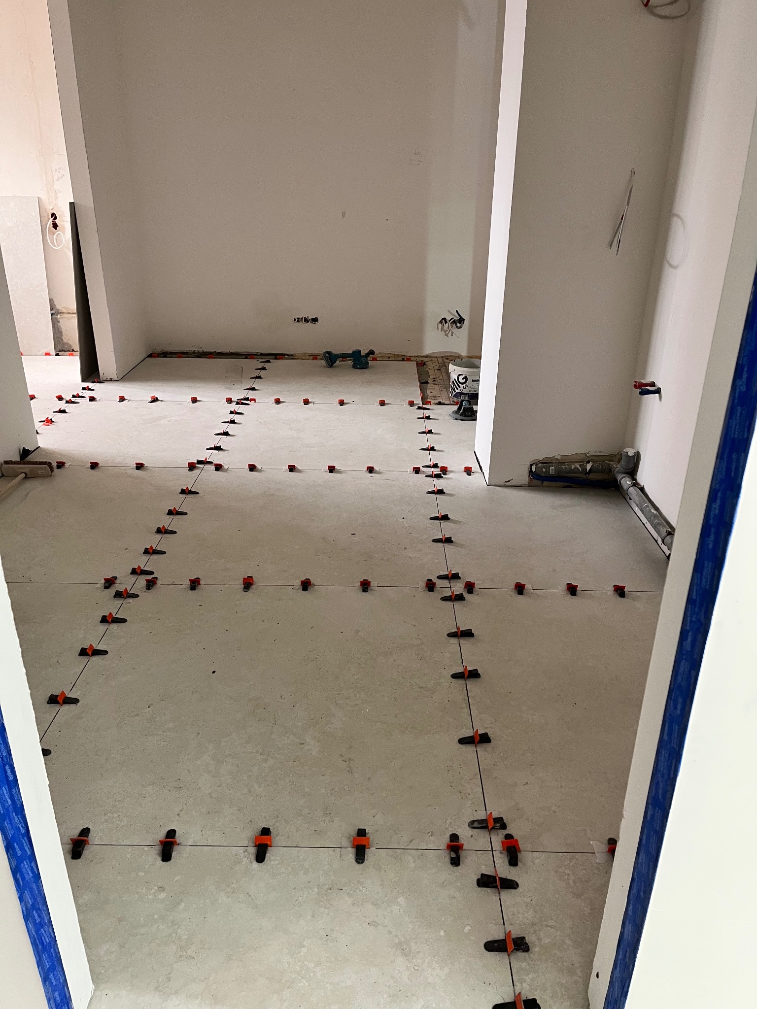 Układanie płytek podłogowych z użyciem systemu poziomowania; widoczne narzędzia i etapy prac wykończeniowych w jasnym wnętrzu.