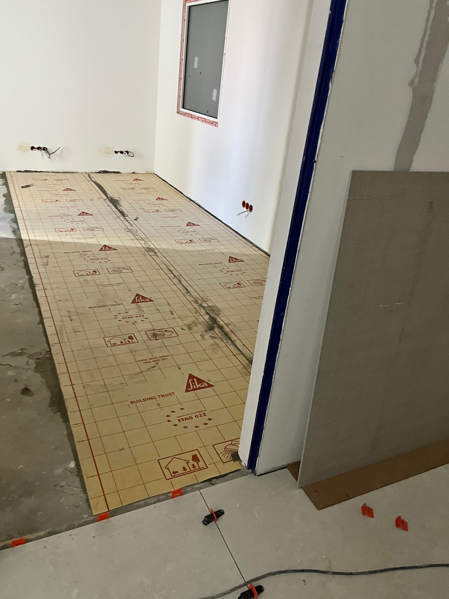 Przygotowanie podłoża pod ogrzewanie podłogowe: rozłożona mata grzewcza Sika z czerwonym nadrukiem na surowym betonie. Widoczne wystające kable i płyta karton-gips.