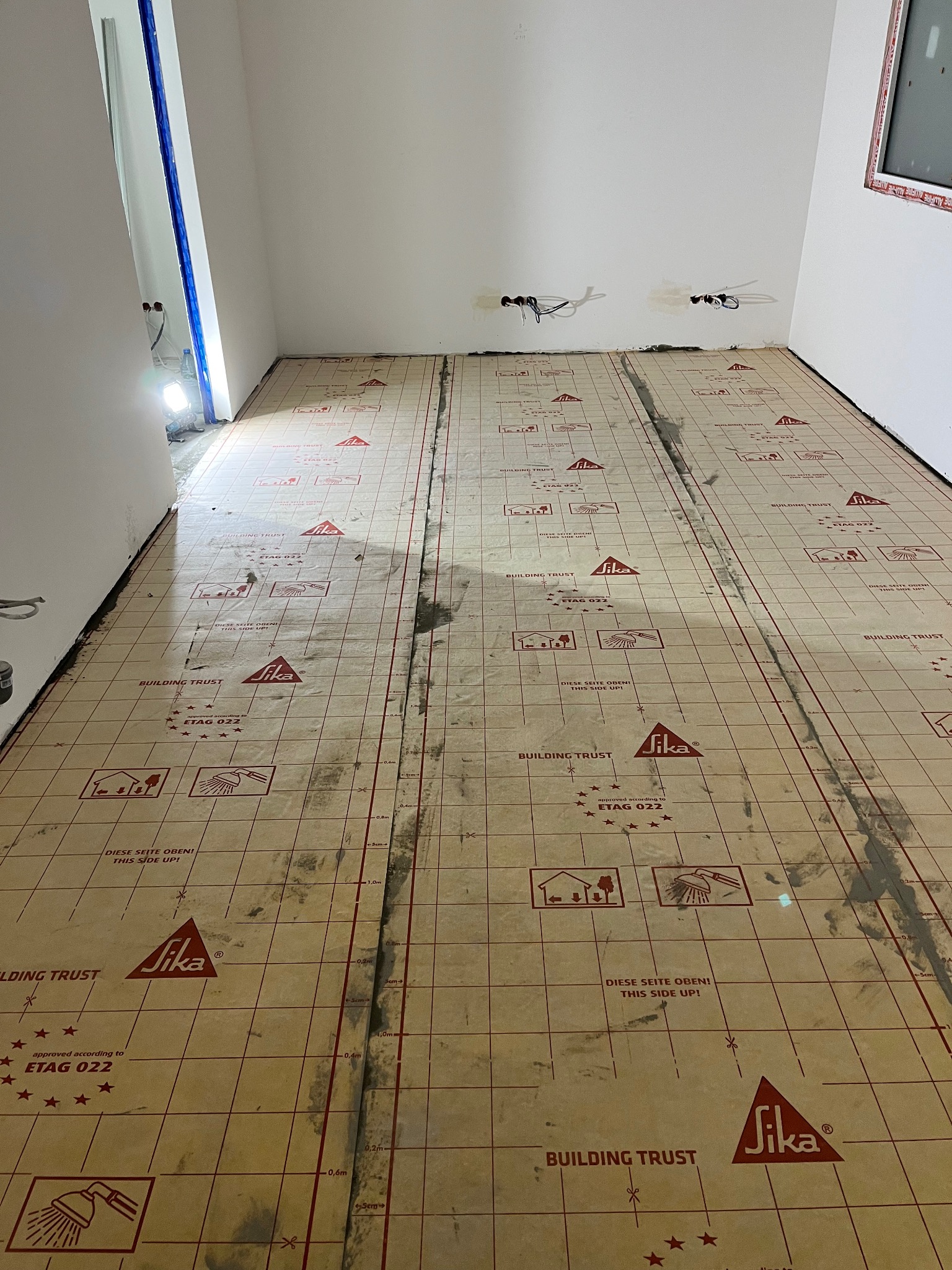 Przygotowana podłoga z izolacją i folią grzewczą JIKA w pomieszczeniu przed wylaniem wylewki, widoczne kable elektryczne wystające ze ściany.