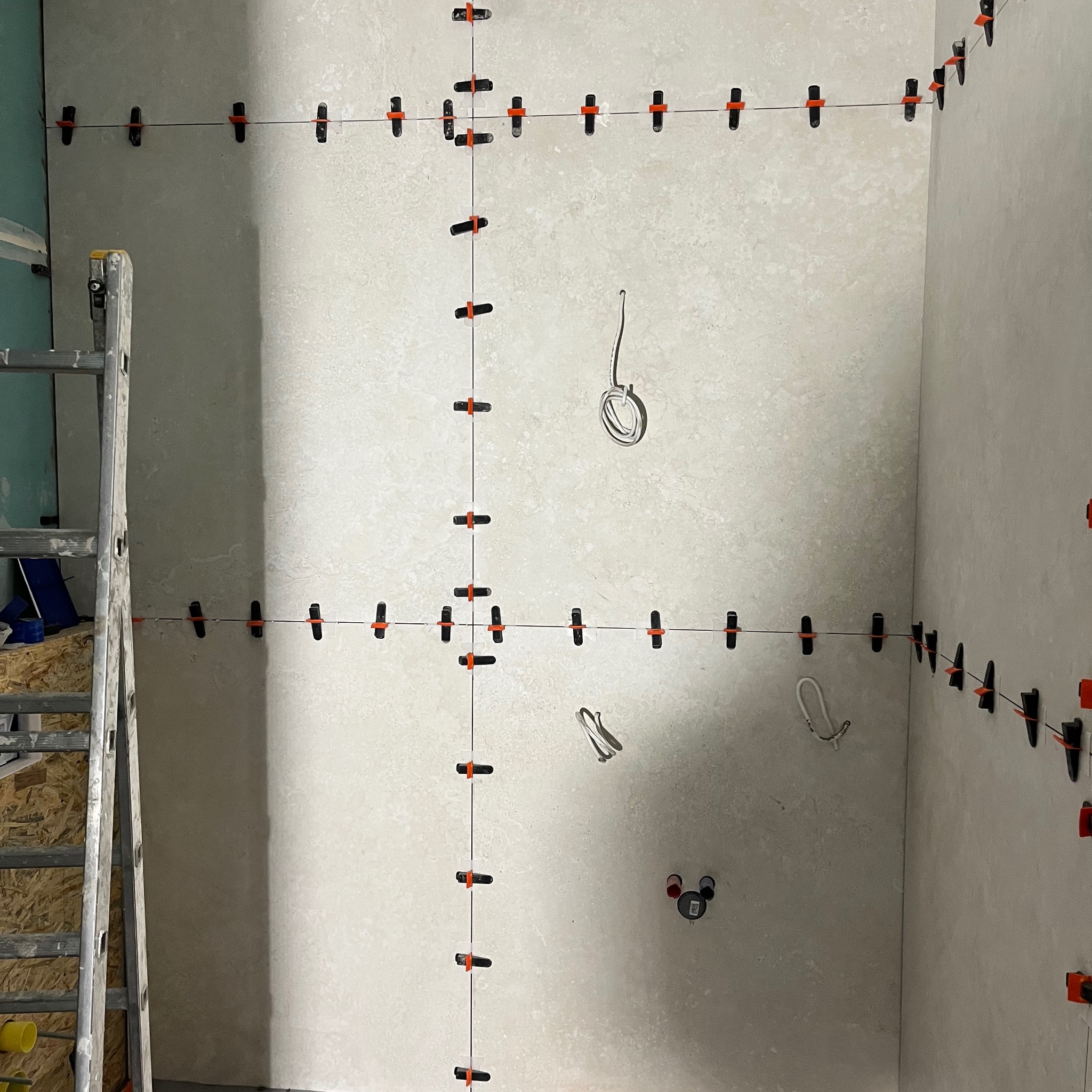 Ściana wyłożona płytkami z użyciem klipsów poziomujących, widoczna drabina.  Etap prac wykończeniowych wewnątrz pomieszczenia z widoczną instalacją elektryczną.