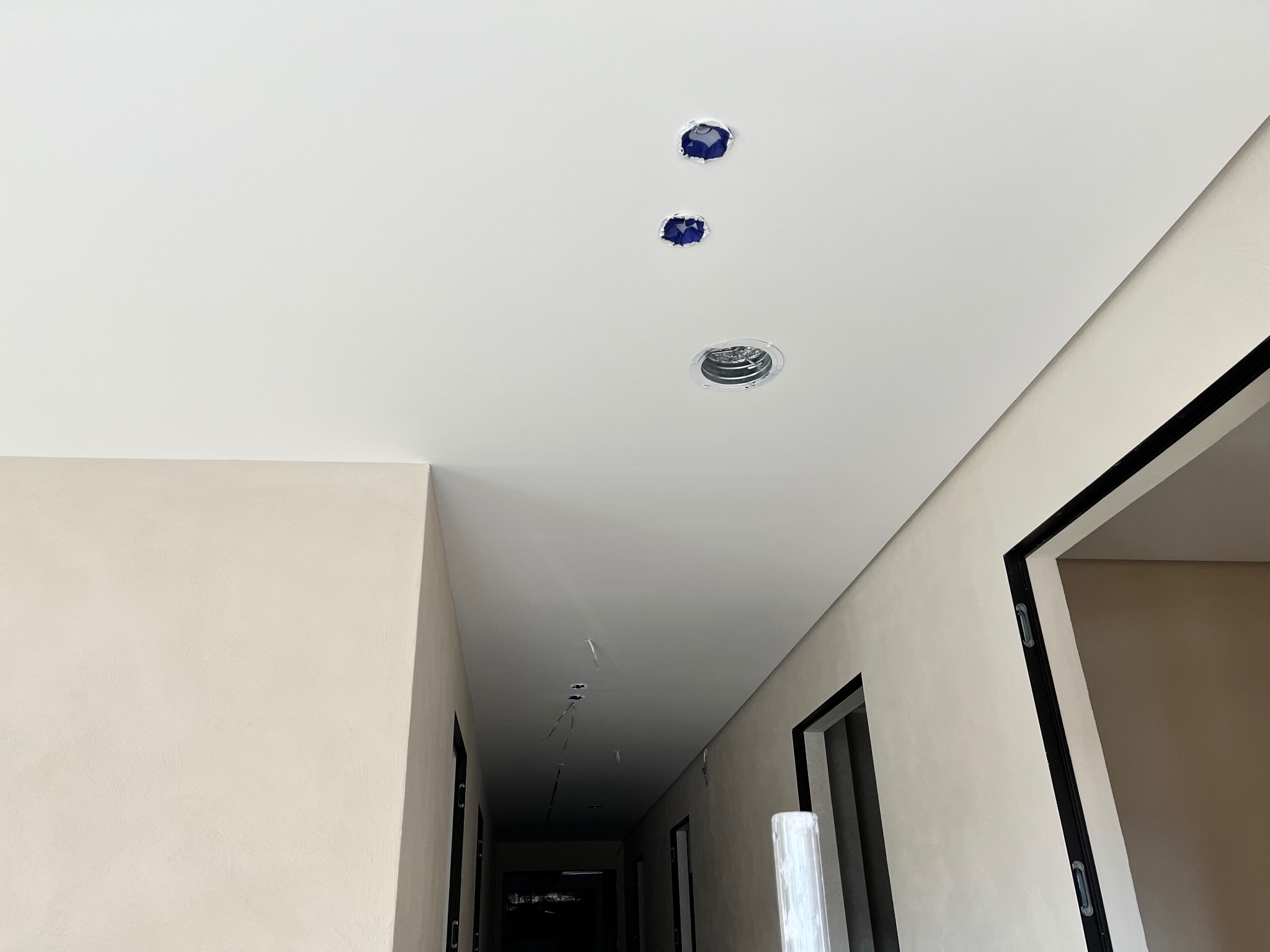 Biały sufit z trzema otworami na oświetlenie, jeden z zamontowaną oprawą, w korytarzu z beżowymi ścianami i czarnymi obramowaniami drzwi.