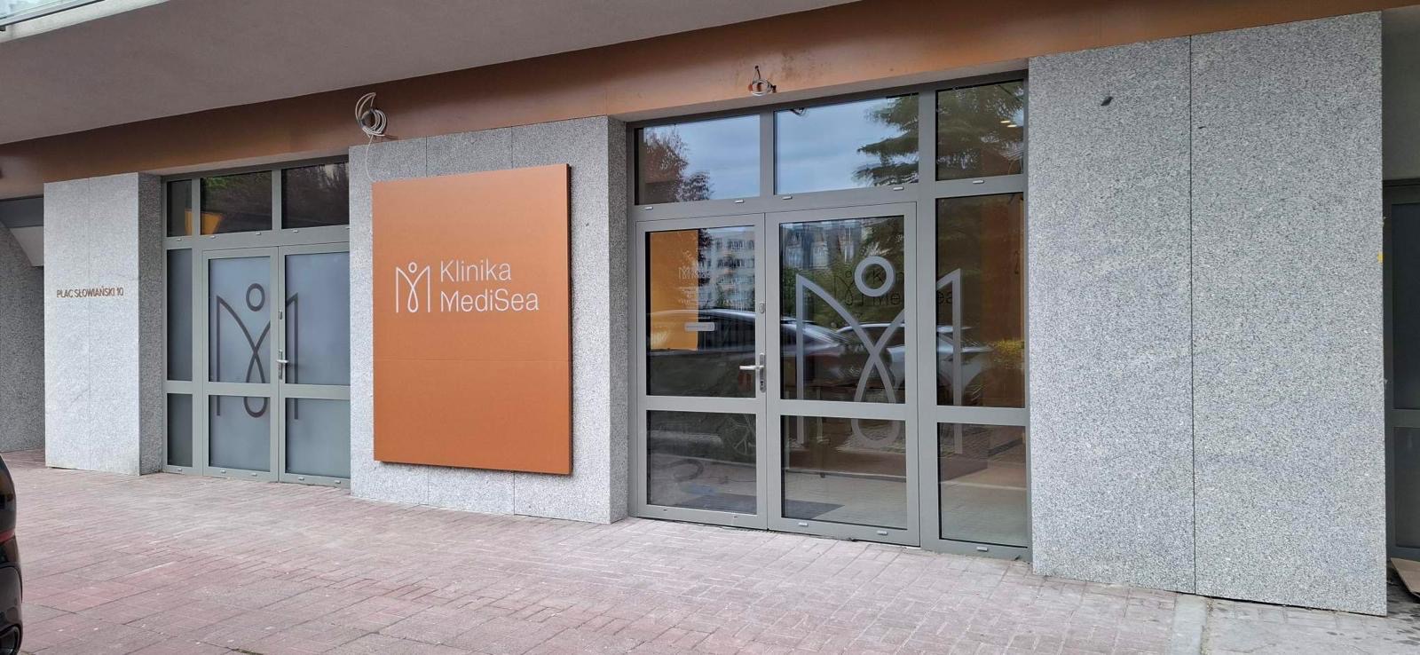 Elewacja budynku z szarego kamienia z dużym, pomarańczowym szyldem „Klinika MediSea”. Wejście z szarymi ramami okiennymi i szklanymi drzwiami.