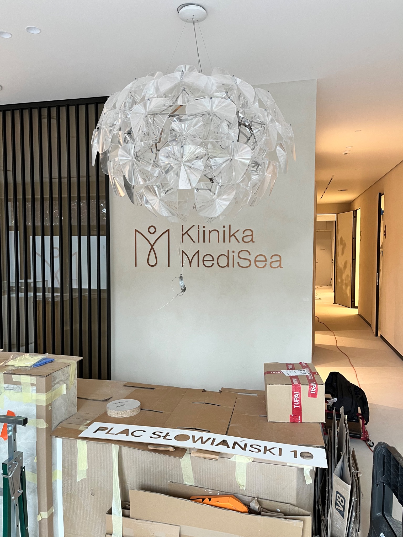Wnętrze w trakcie wykańczania: lampa nad kartonami i szablonem napisu 'Plac Słowiański 10', logo 'Klinika MediSea' na ścianie, widok na korytarz w tle.