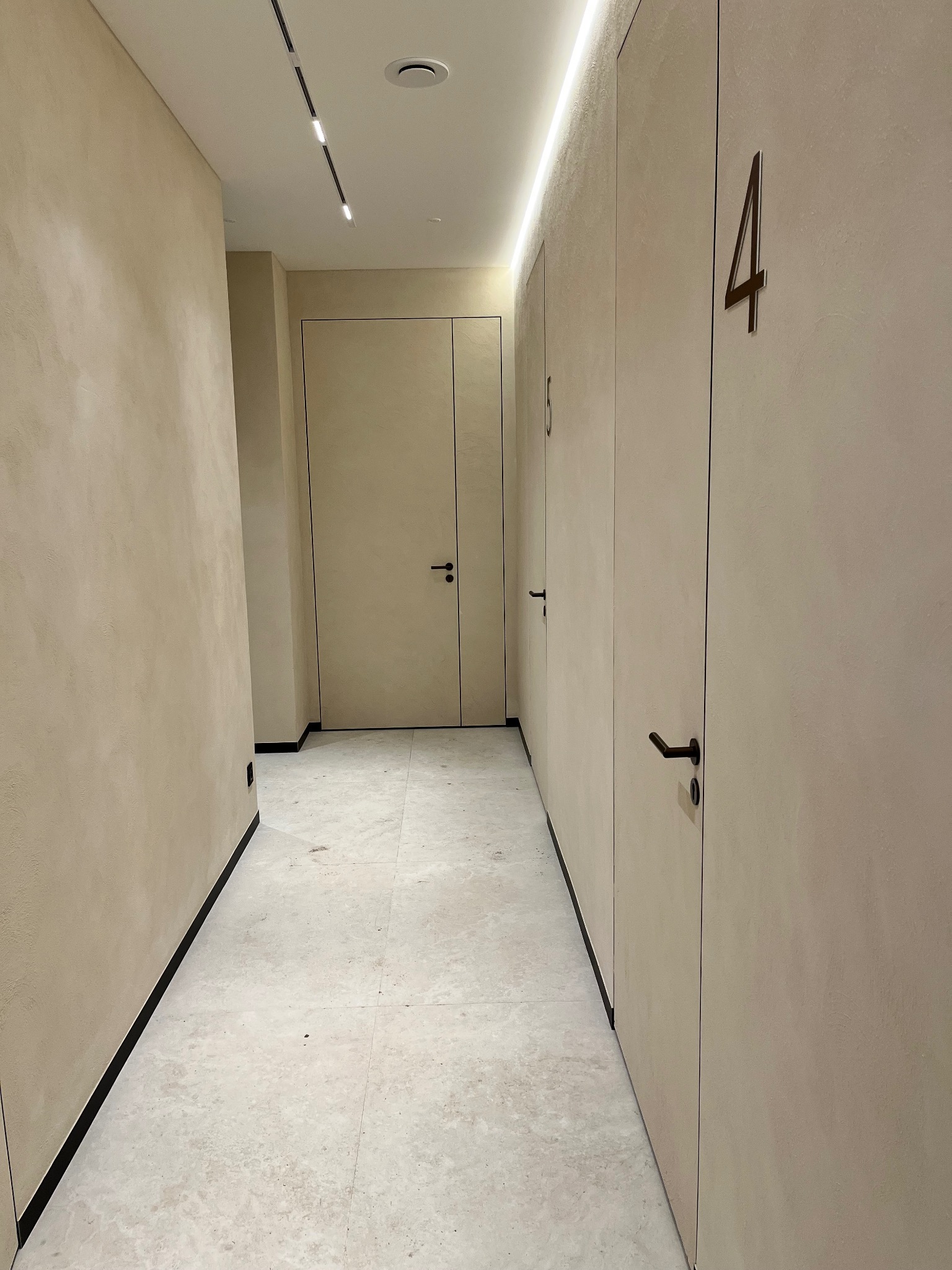 Korytarz w neutralnych barwach z numerami na drzwiach, oświetleniem ledowym i wykończeniem ścian imitującym tynk strukturalny. Widoczne minimalistyczne detale.