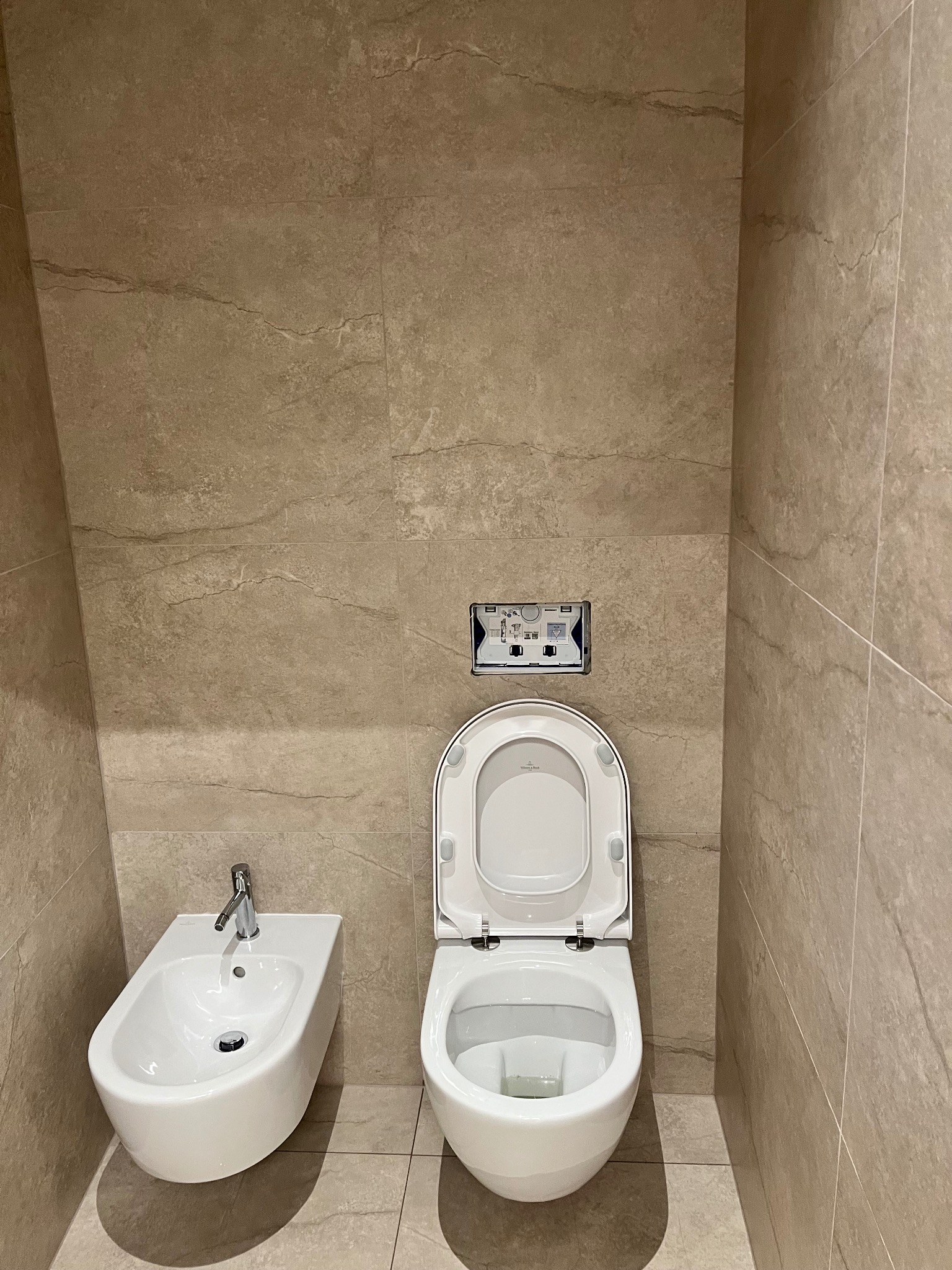 Beżowa łazienka z płytkami imitującymi kamień: biała toaleta z otwartą deską i bidet, chromowana armatura, minimalistyczny design.