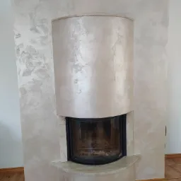 Otynkowany kominek z dekoracyjną strukturą w odcieniach beżu i szarości, z widocznymi refleksami światła, na tle drewnianej podłogi.