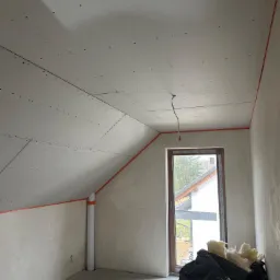 Poddasze w trakcie wykańczania: zamontowane płyty kartonowo-gipsowe na skosach sufitu, widoczne profile metalowe na podłodze, okno z widokiem na zewnątrz i worki z wełną mineralną w rogu...