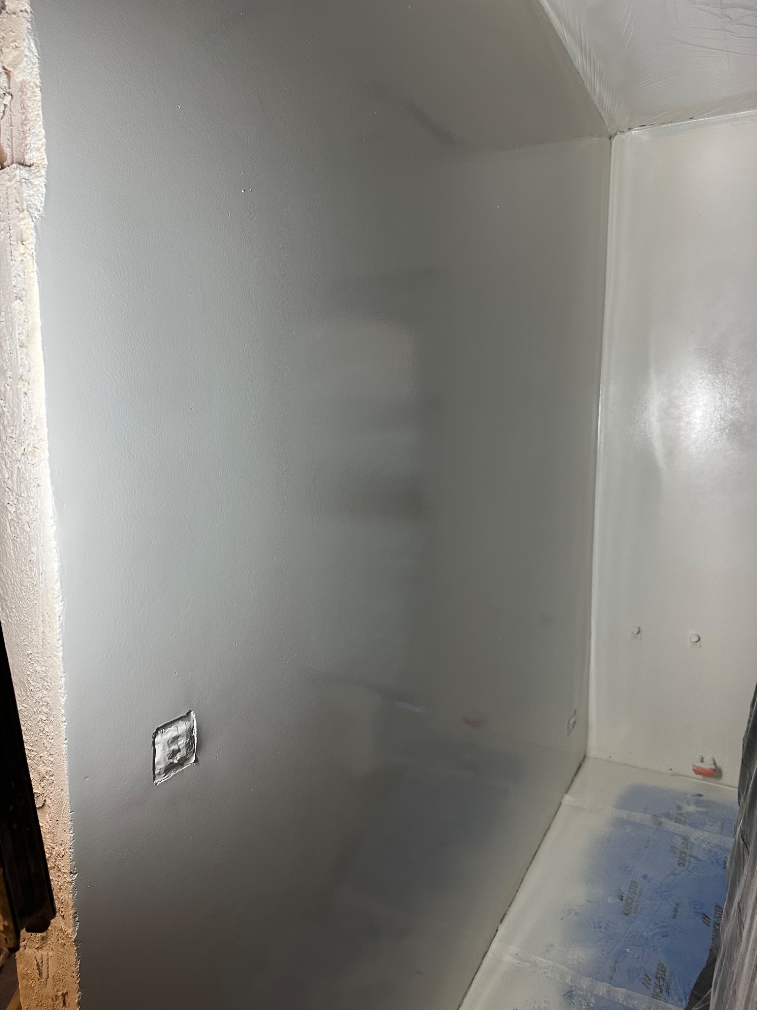 Świeżo pomalowane na szaro ściany w pomieszczeniu, widoczne zabezpieczenie podłogi folią, surowy narożnik ściany i puszka elektryczna bez osprzętu.