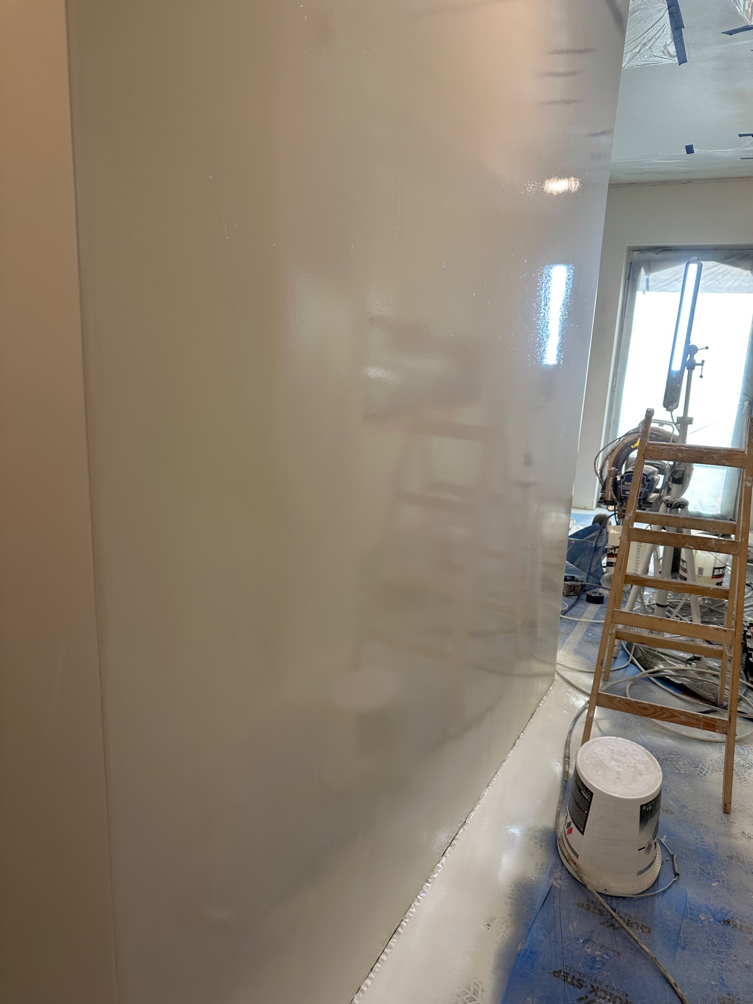 Świeżo pomalowana ściana w jasnym odcieniu z połyskiem, widoczna drabina, wiadro z farbą oraz maszyna do malowania natryskowego w tle, zabezpieczona podłoga.