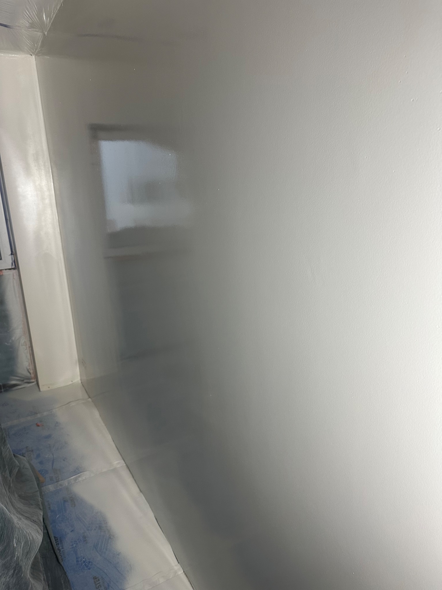 Świeżo pomalowana, gładka, szara ściana w rogu pomieszczenia, zabezpieczona folią podłoga, widoczne okno w odbiciu.