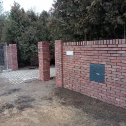 Nowo wymurowany, ceglany mur ogrodzeniowy z bramą wejściową i skrzynką gazową, otoczony ziemią i zielenią.