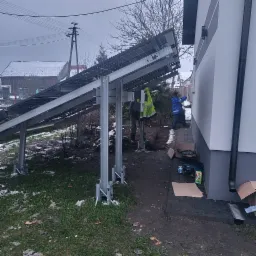 Instalacja naziemnej konstrukcji pod panele słoneczne przy ścianie budynku; widoczny pracownik w niebieskiej kurtce montujący elementy na ścianie, obok skrzynki z narzędziami i kartony.