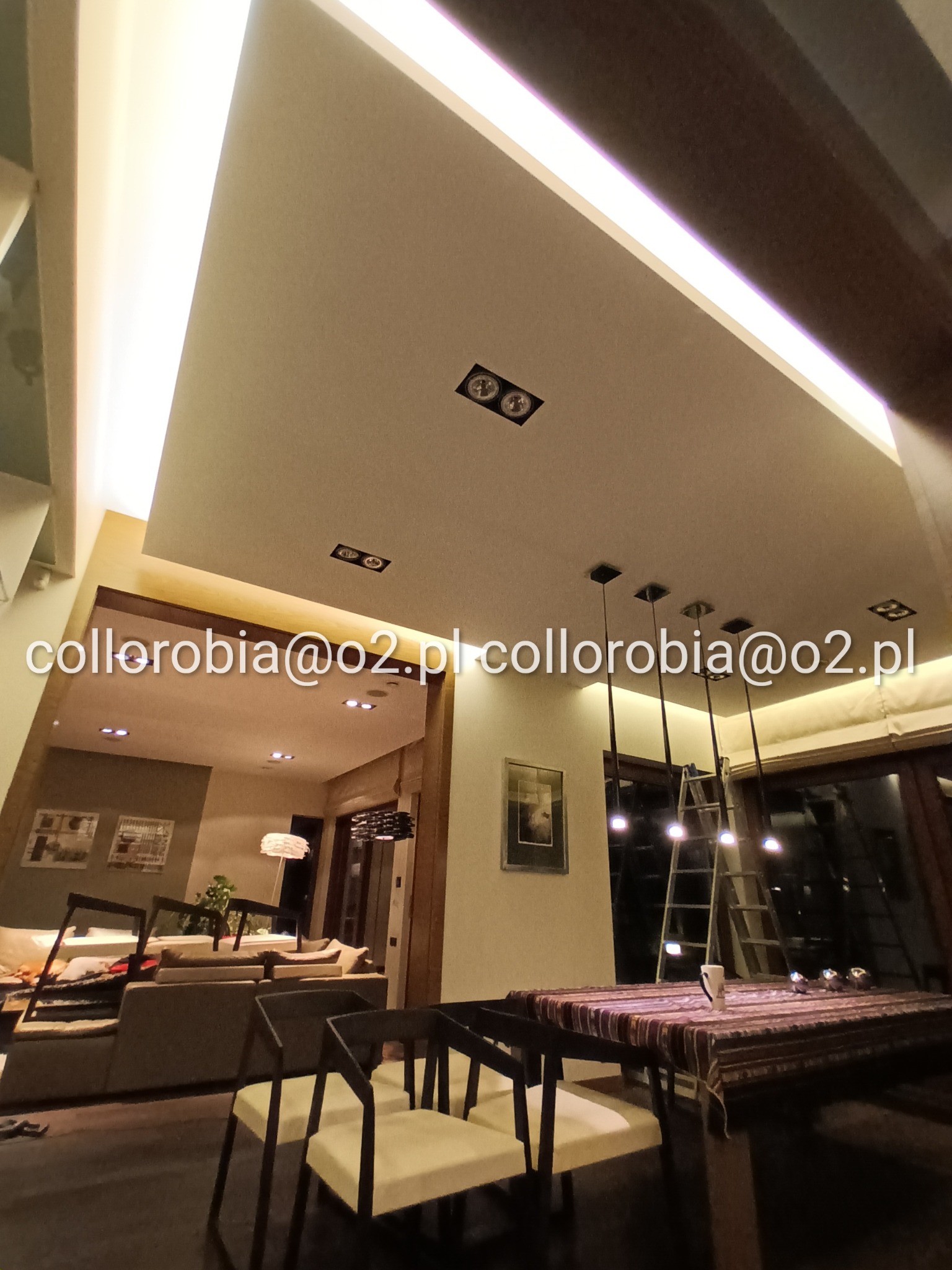 Nowoczesne oświetlenie LED w suficie podwieszanym, widok na jadalnię z ciemnymi krzesłami i stołem z wzorzystym obrusem, w tle salon z sofą i dekoracjami ściennymi, drabina oparta o okno.