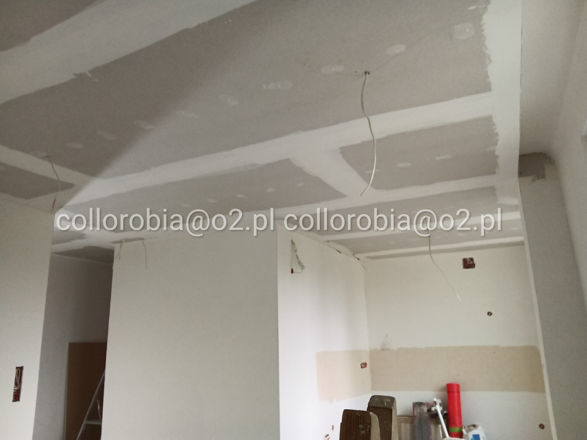 Wnętrze w trakcie wykańczania: widoczne płyty gipsowo-kartonowe na suficie i ścianach, częściowo zaszpachlowane, z wystającymi przewodami elektrycznymi. Na ścianie widoczne ślady po instalacjach...