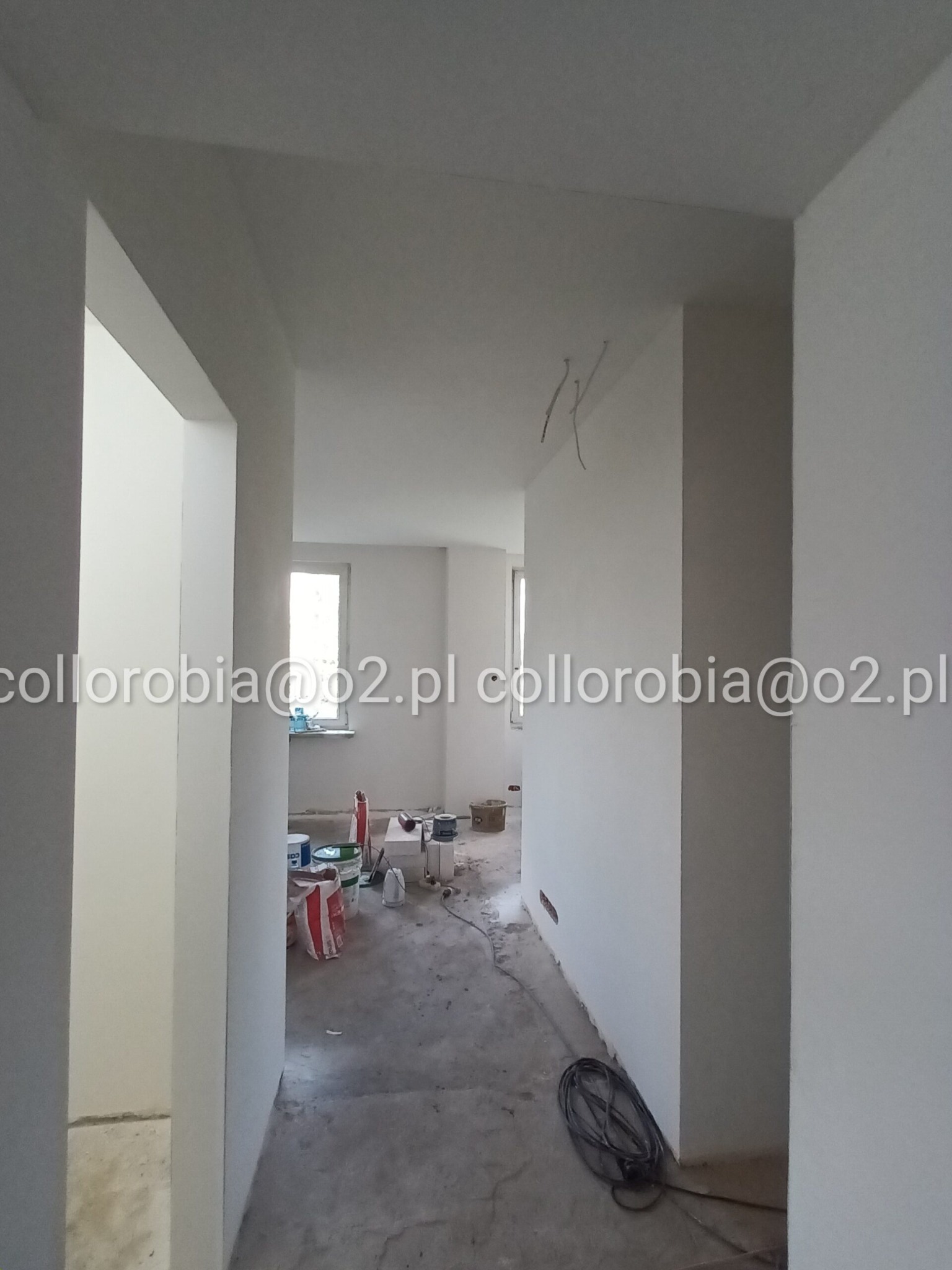Korytarz w trakcie prac wykończeniowych, świeżo pomalowane ściany na biało, widoczne kable wystające ze ściany, worki z gładzią i narzędzia malarskie w tle, podłoga przygotowana pod dalsze prace.