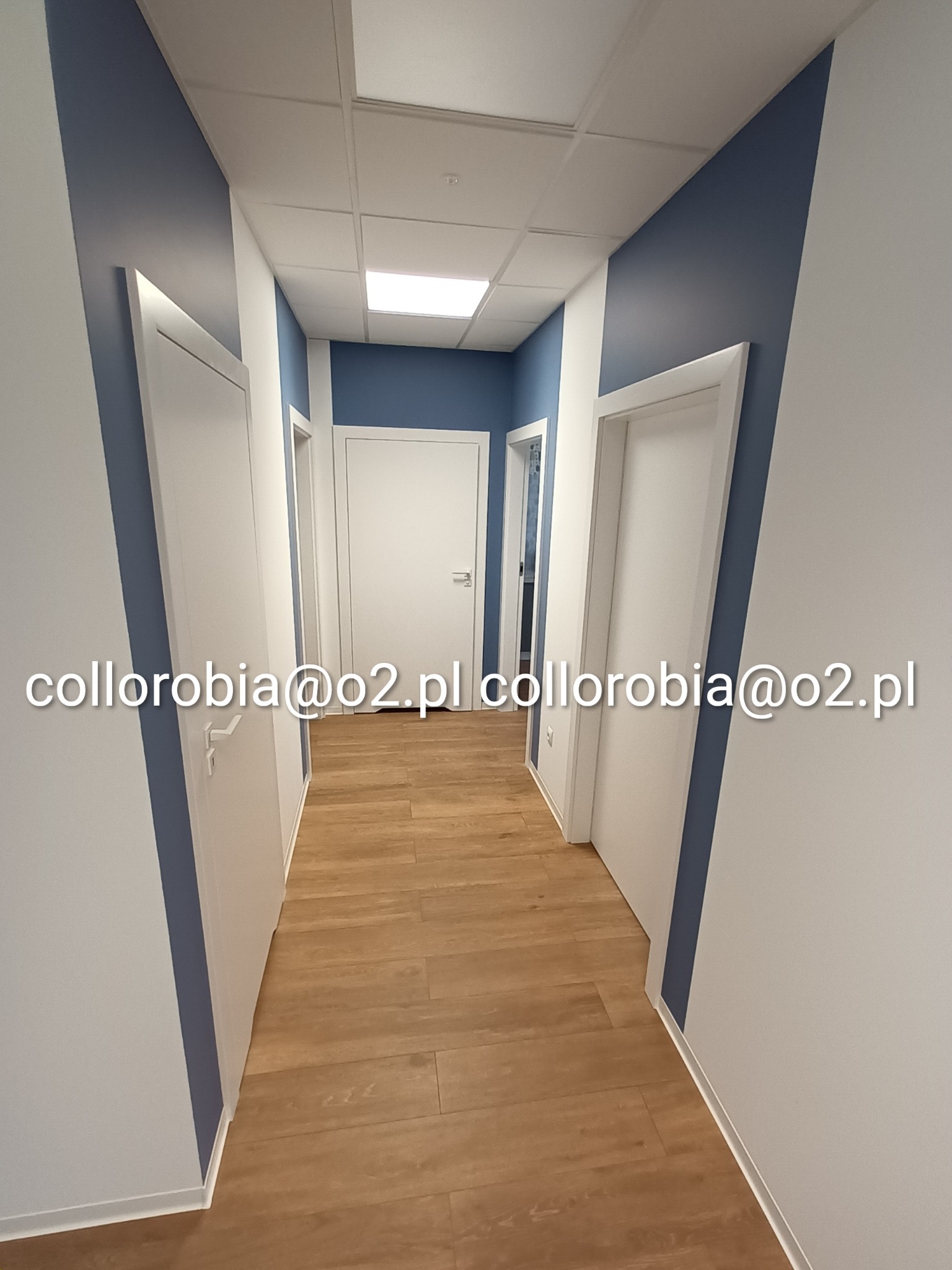 Wnętrze lokalu usługowego po remoncie: korytarz z drewnopodobną podłogą, białe ściany z pionowymi pasami w kolorze granatowym, białe drzwi i sufit z oświetleniem.