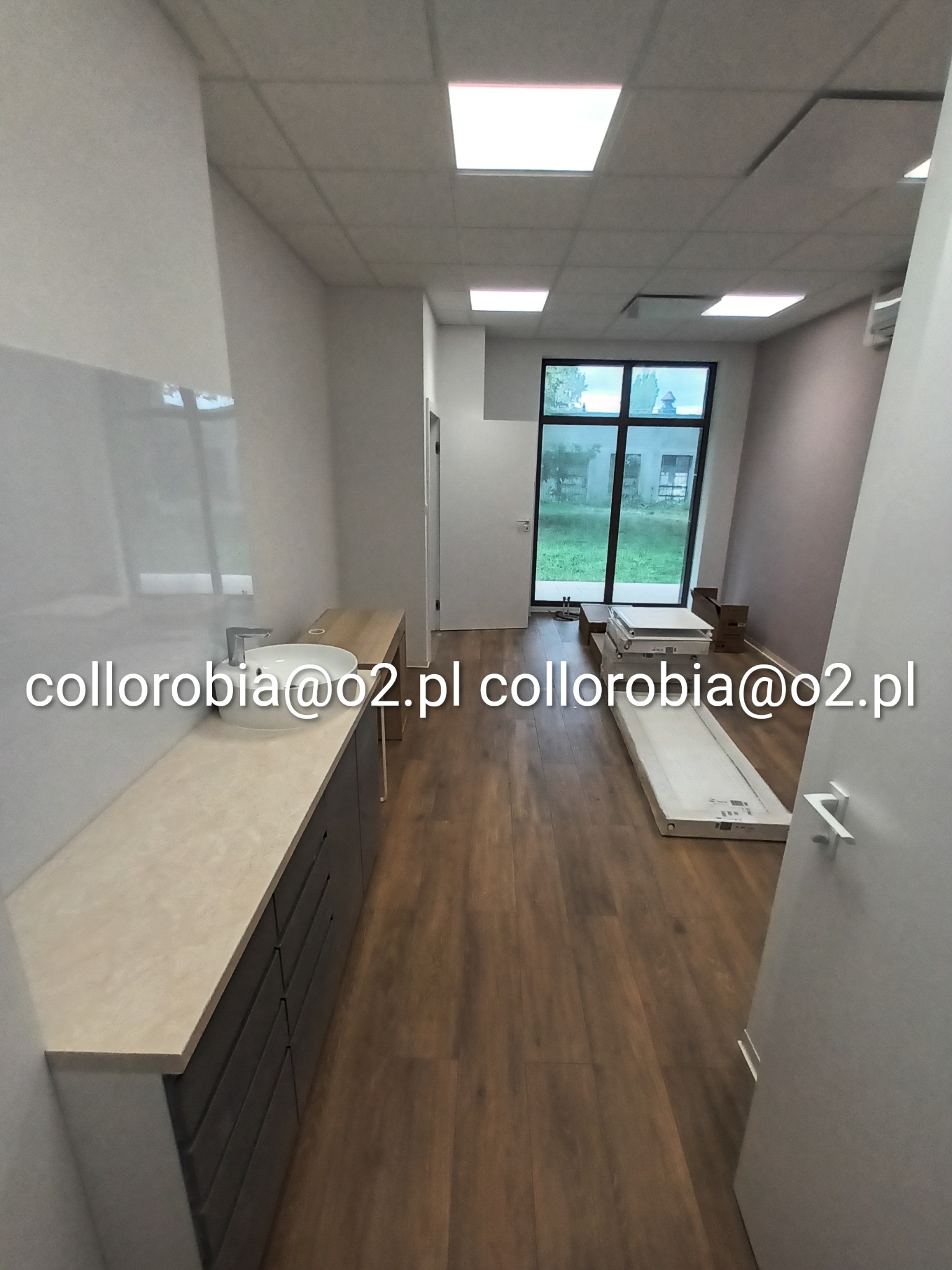 Nowe, jasne pomieszczenie usługowe z umywalką w zabudowie, świeżo położoną podłogą drewnianą i elementami do wykończenia wnętrza przy oknie.