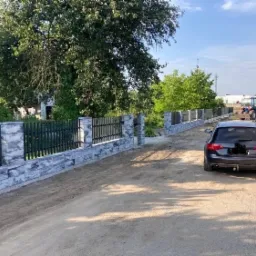 Nowoczesne ogrodzenie z bloczków Joniec o szarej, marmurkowej fakturze, z czarnymi, metalowymi przęsłami, w trakcie prac ziemnych na nierównym terenie. W tle widoczny traktor i samochód dostawczy.