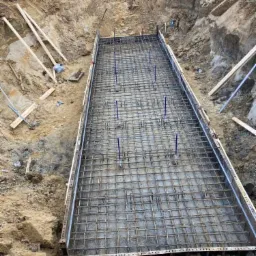 Szeroki wykop z przygotowanym zbrojeniem w metalowej szalunku, widoczne wystające pręty pionowe, ziemia wokół wykopu, drewniane podpory i taśma ostrzegawcza w tle.