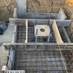 Zbrojenie fundamentu w wykopie z drewnianym szalunkiem, widoczne stalowe pręty i elementy konstrukcyjne, ziemia wokół wykopu.