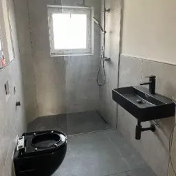 Nowoczesna łazienka w szarościach z czarną armaturą: podwieszana toaleta, umywalka nablatowa i kabina prysznicowa typu walk-in z czarnym zestawem natryskowym.