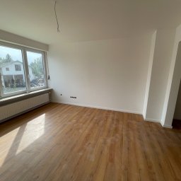 Krawbud Monika Sosnowska - Jasne, przestronne wnętrze po remoncie z drewnopodobną podłogą, białymi ścianami i dużym oknem z widokiem na dom i zieleń. Widoczne ślady po remoncie na podłodze i wystające przewody elektryczne...