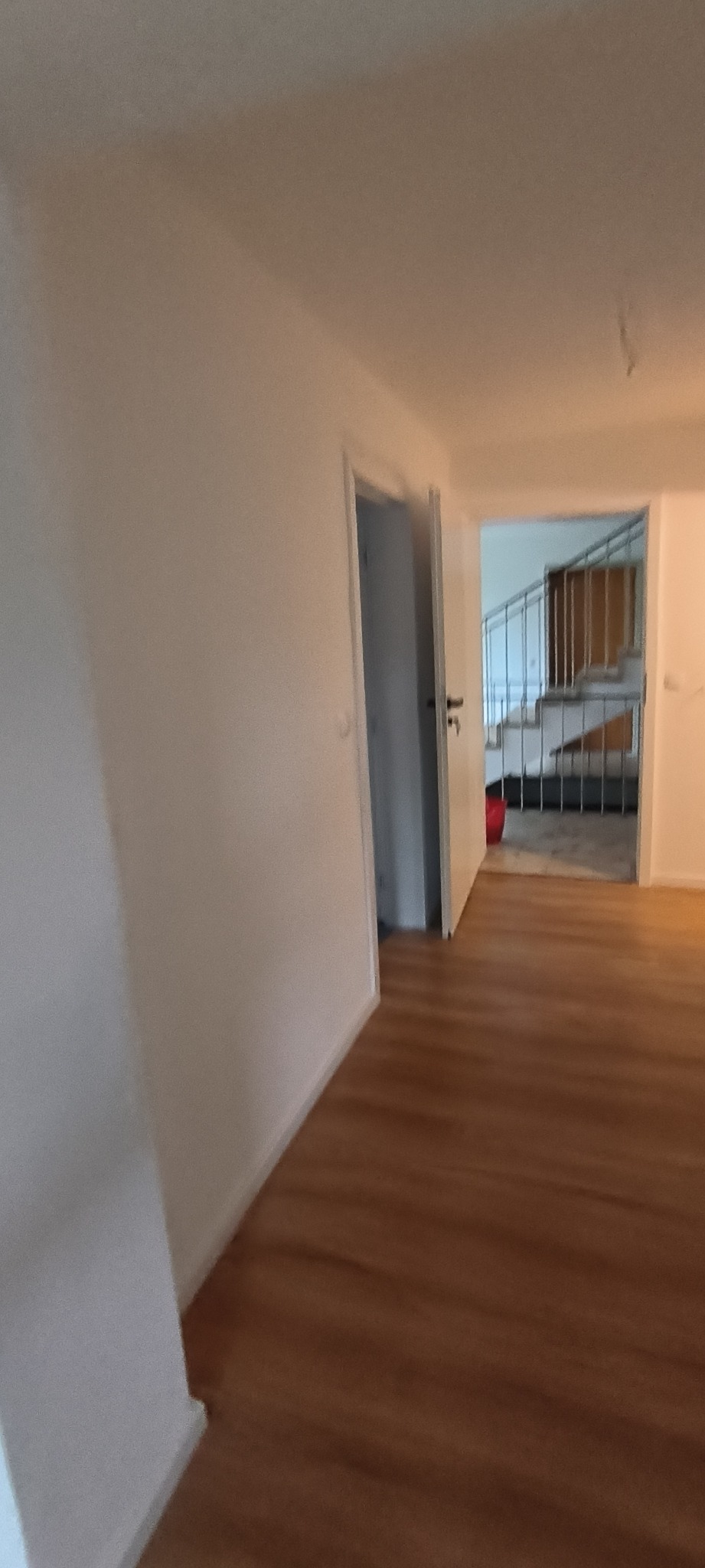 Wnętrze mieszkania po remoncie: widok na korytarz z drewnianą podłogą, białe ściany, otwarte drzwi do pokoju z schodami i metalową barierką.