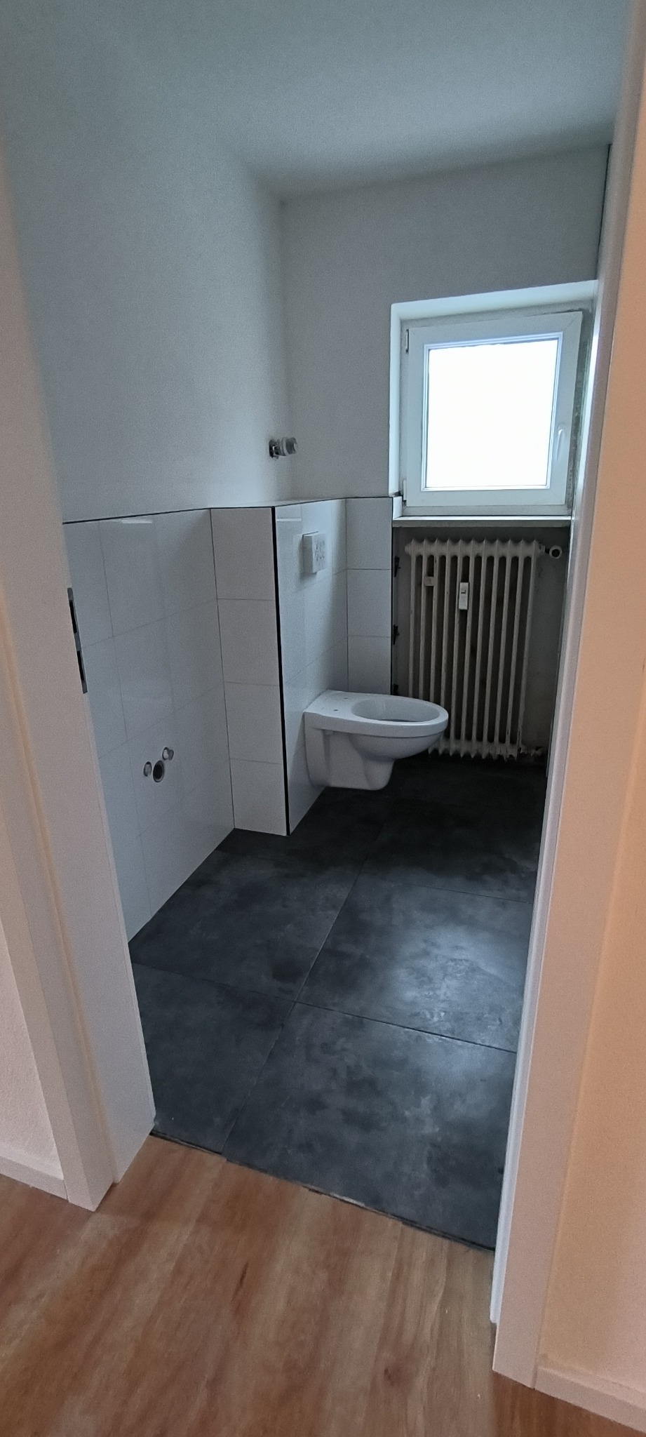 Wnętrze małej, jasnej łazienki w trakcie prac wykończeniowych, widoczne białe płytki na ścianach, ciemnoszara podłoga, wisząca toaleta bez deski i grzejnik pod oknem, brak elementów dekoracyjnych.
