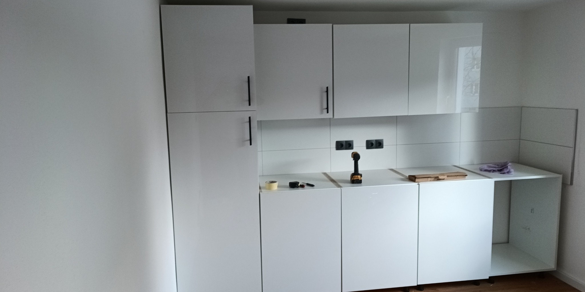 Montaż nowoczesnej, białej kuchni na wymiar: widoczne szafki wiszące i stojące, narzędzia (wiertarka) na blacie, płytki na ścianie, jeszcze bez sprzętu AGD.