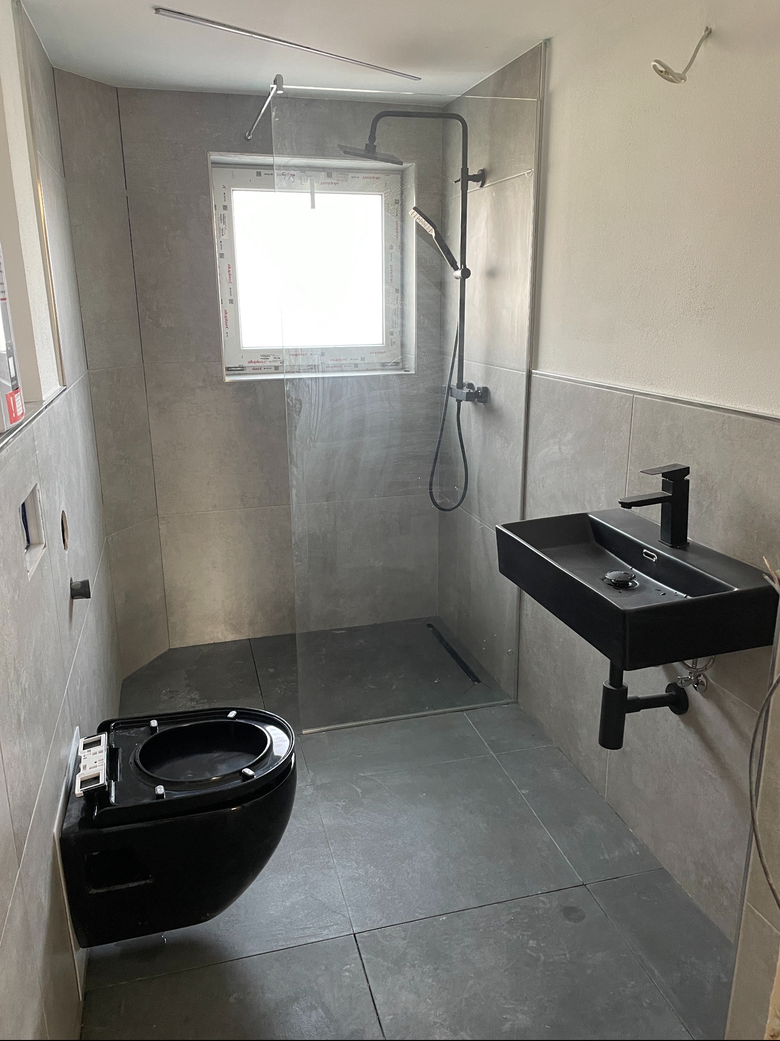 Nowoczesna łazienka w szarościach z czarną armaturą: podwieszana toaleta, umywalka nablatowa i kabina prysznicowa typu walk-in z czarnym zestawem natryskowym.