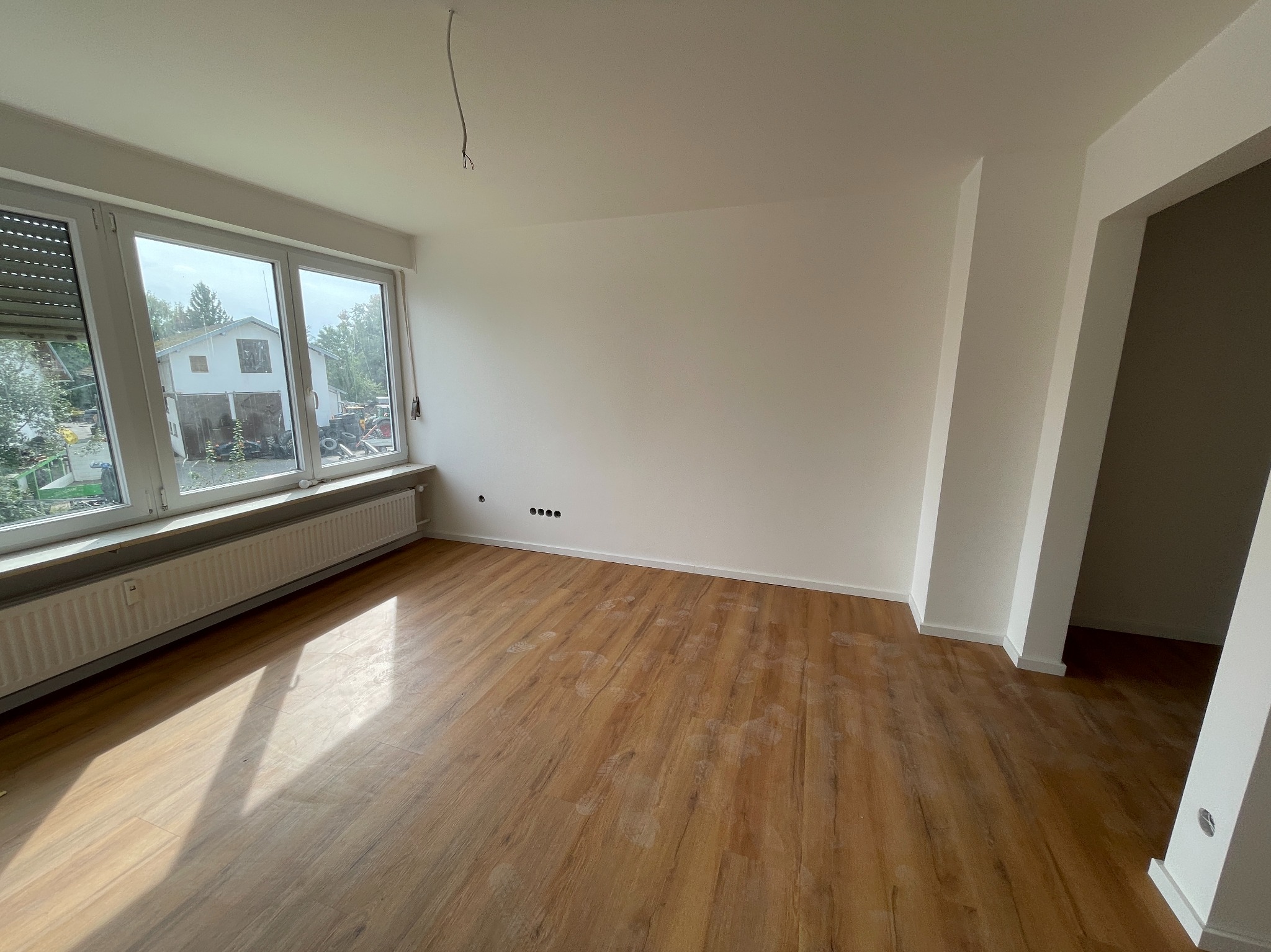 Jasne, przestronne wnętrze po remoncie z drewnopodobną podłogą, białymi ścianami i dużym oknem z widokiem na dom i zieleń. Widoczne ślady po remoncie na podłodze i wystające przewody elektryczne...