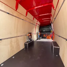 Wnętrze pustego, wyłożonego sklejką i zabezpieczonego szynami transportowymi, czerwonego busa dostawczego, przygotowanego do transportu ładunków.