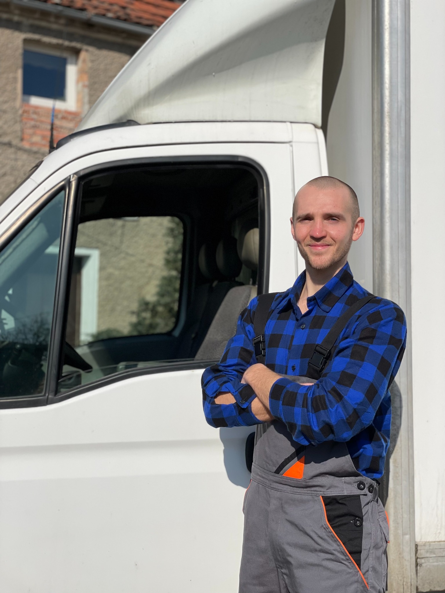 Uśmiechnięty mężczyzna w niebiesko-czarnej koszuli w kratę i szarych ogrodniczkach z pomarańczowymi wstawkami, stojący na zewnątrz obok białego samochodu dostawczego z budynkiem w tle.